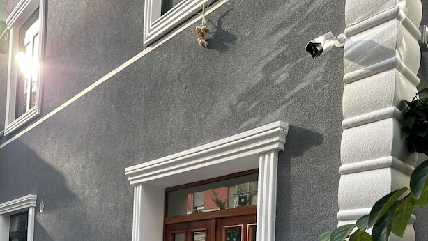 Fachada de edificio con pared gris texturizada, puerta de madera con paneles de vidrio y un columnilla decorativa blanca.