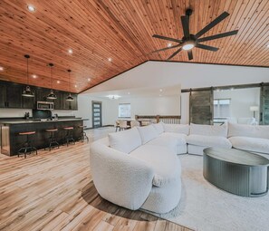 Een ruime woonkamer met een houten plafond, een grote witte modulaire bank, een ronde salontafel en een keuken met moderne apparatuur.