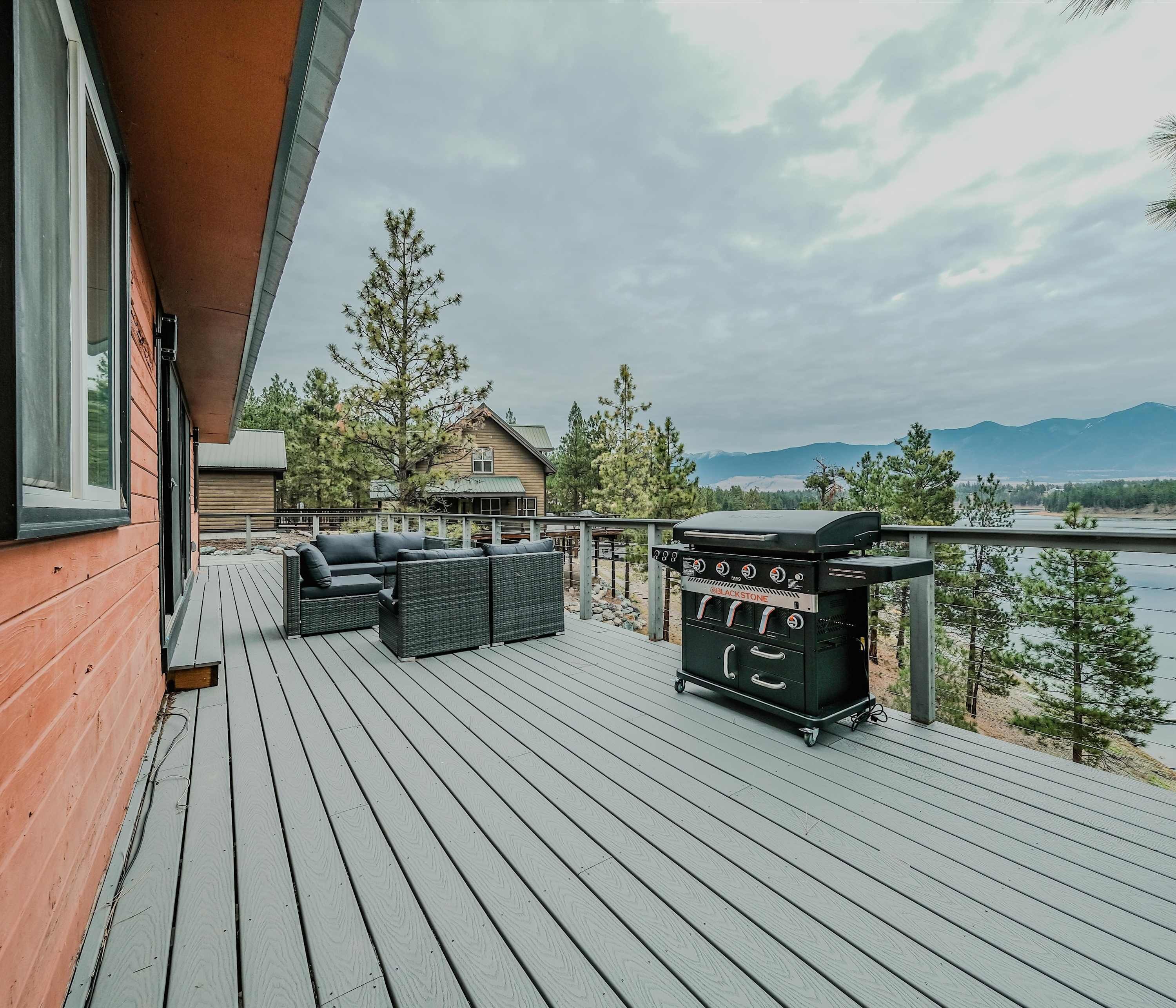 Eine Holzterrasse mit Gartenmöbeln und einem Grill, mit Blick auf einen See und Berge.