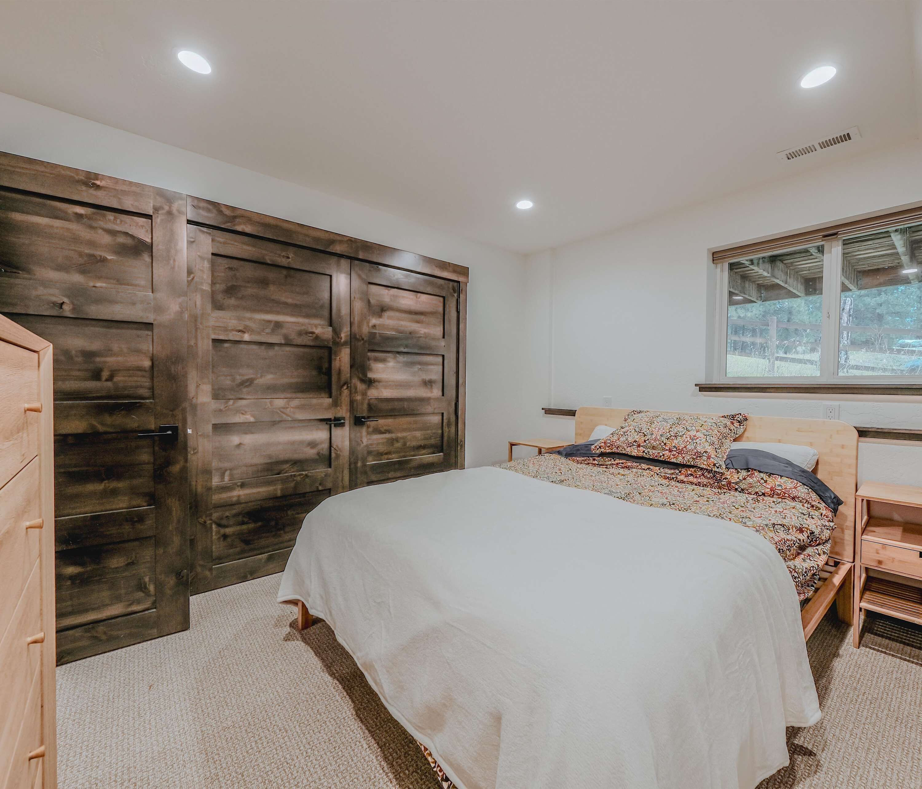 Un dormitorio con un armario de madera grande, una cama con ropa de cama estampada, una mesita de noche pequeña y una ventana con vistas a zonas verdes.
