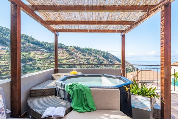 A balcony with a hot tub, wicker furniture, and a view of a hilly landscape.
