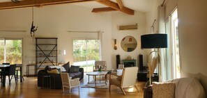 Ein geräumiges Wohnzimmer mit Holzboden, einem schwarzen Sofa, einem weißen Sessel, einem runden Couchtisch und einem Fernseher. Ein Bücherregal, ein Spiegel und ein Fenster mit Vorhängen sind ebenfalls vorhanden.