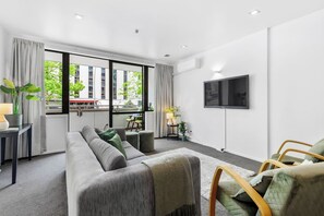 Habitación de hotel moderna con sofá gris, una mesita, televisión montada en la pared y ventanas amplias con vistas a la ciudad.