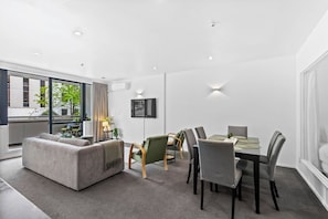 A modern living room with a sofa, dining table, chairs, and a large window offering a view of the outdoors. - City Centre Living Meets Comfort by Zodiak (Auckland)