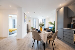 A modern dining area with a table set for four, a kitchen area with grey cabinets, and a view of a bedroom with a yellow blanket. - Highrise Heaven 2BR | Manchester City Centre (Manchester)