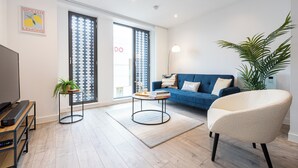 Ein modernes Wohnzimmer mit einem blauen Sofa, einem weißen Sessel, einem Couchtisch, einem Fernseher und einem großen Fenster mit Blick auf die Stadt.