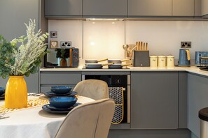 A modern kitchen with grey cabinets, a white countertop, and a dining area with a table set for two. - Highrise Heaven 2BR | Manchester City Centre (Manchester)