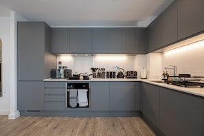A modern kitchen with grey cabinets, a built-in oven, and a countertop with various appliances and utensils. - Royal Ebony | Luxe 2BR City Centre Apartment (Manchester)
