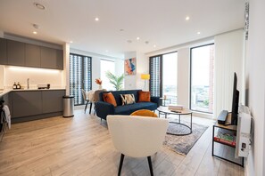 En moderne stue med sofa, sofabord og fjernsyn. Der er et køkkenområde med skabe og vask. Store vinduer giver naturligt lys og udsigt over byen.
