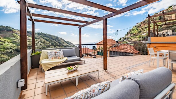 Ein überdachter Außenbereich mit einer Couch, einem Tisch und Stühlen mit Blick auf einen Hügel und Häuser.