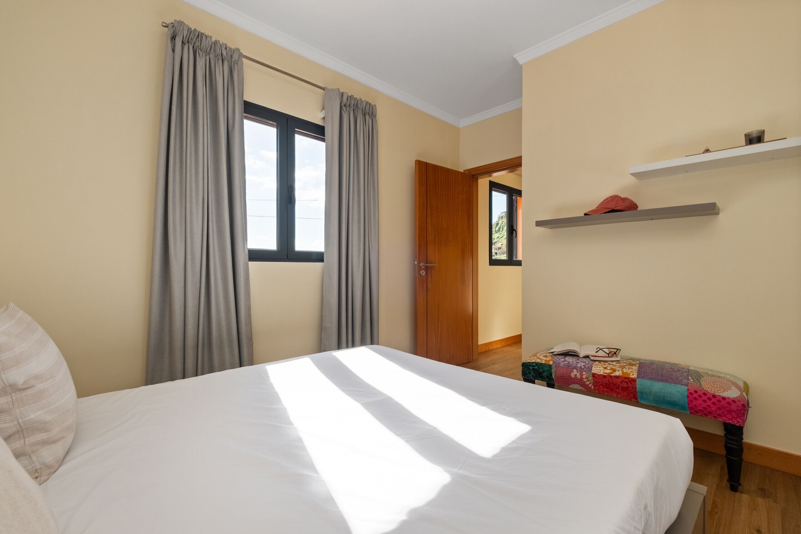 Een slaapkamer met een groot bed, grijze gordijnen, een houten deur en een klein bankje met een sprei.