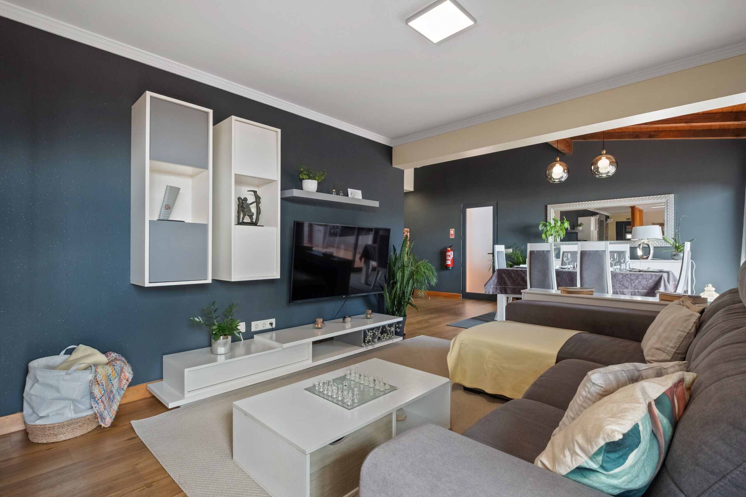 A modern living room with a flat-screen TV, white shelving, a sofa, and a dining area with a mirror and plants.