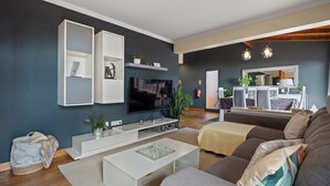 Ein modernes Wohnzimmer mit Flachbildfernseher, weißen Regalen, einer Couch und einem Essbereich mit Spiegel und Pflanzen.