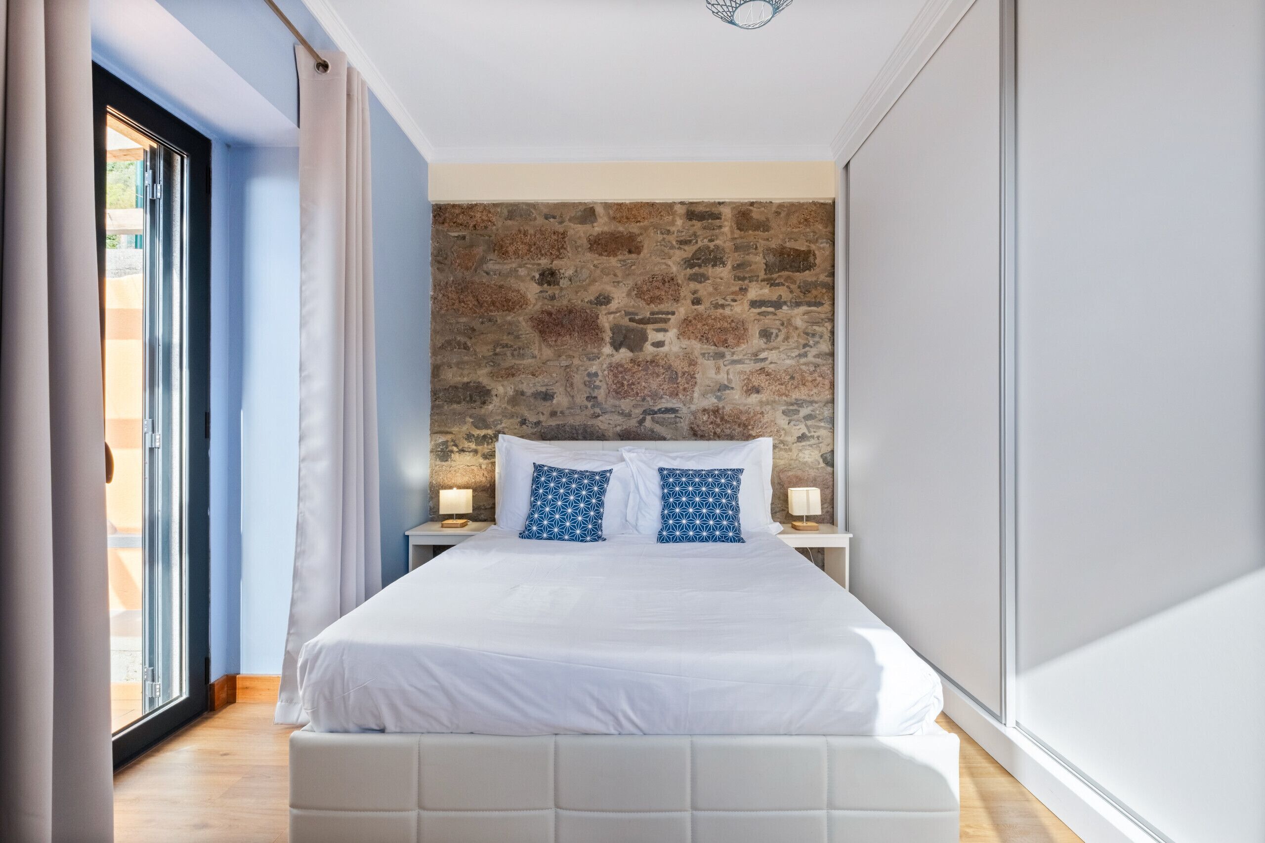 Un dormitorio con pared de piedra, una cama grande con ropa de cama blanca, dos mesitas de noche con lámparas y vistas a través de un ventanal.