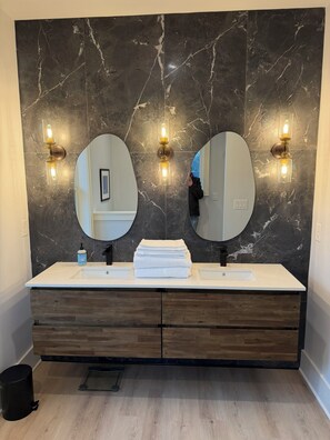 Une salle de bain avec un pare-feu en marbre, deux miroirs ronds, un lavabo blanc et des meubles en bois.