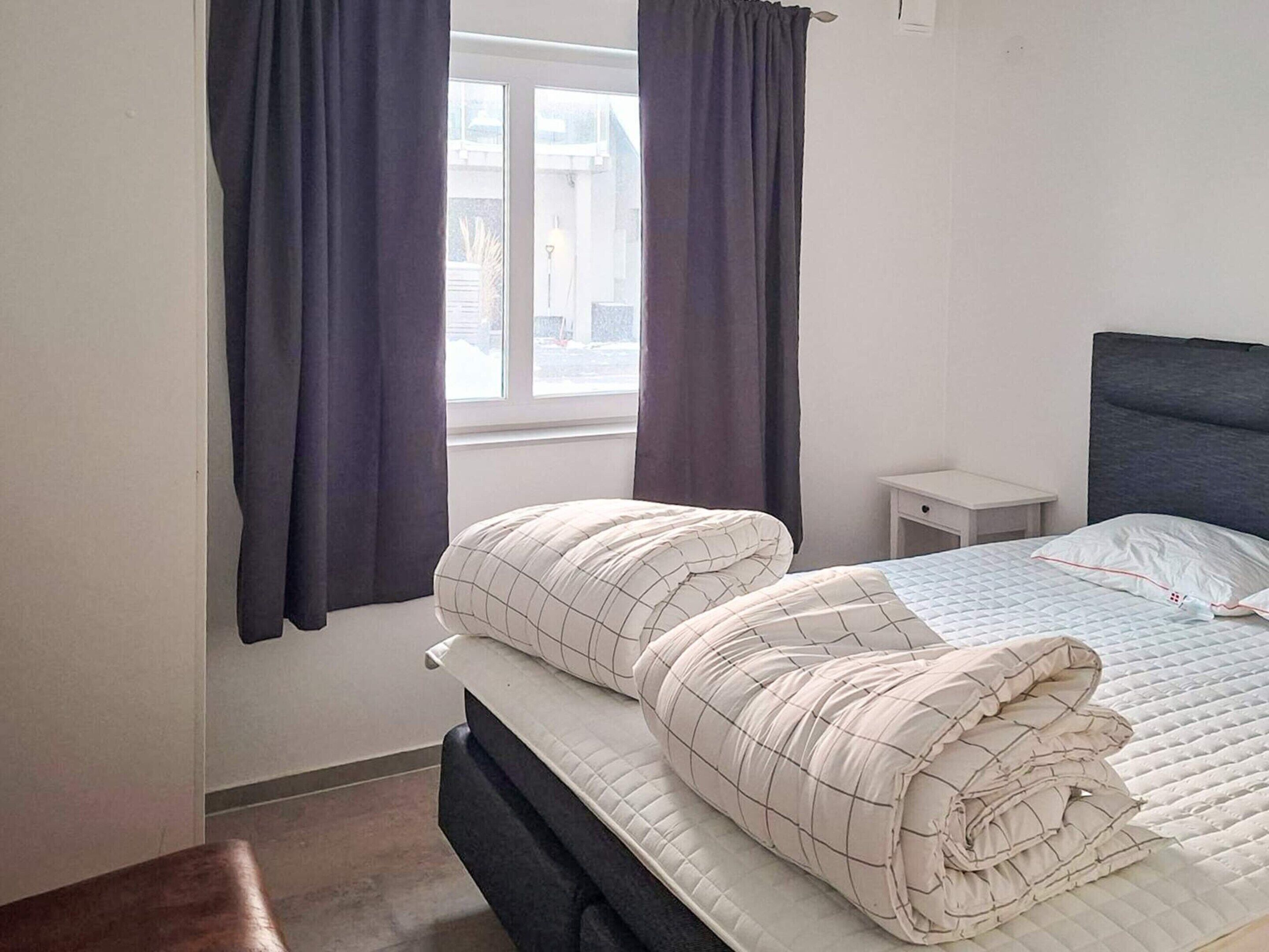 A bedroom with a bed, a nightstand, a window with curtains, and a view of the outside.