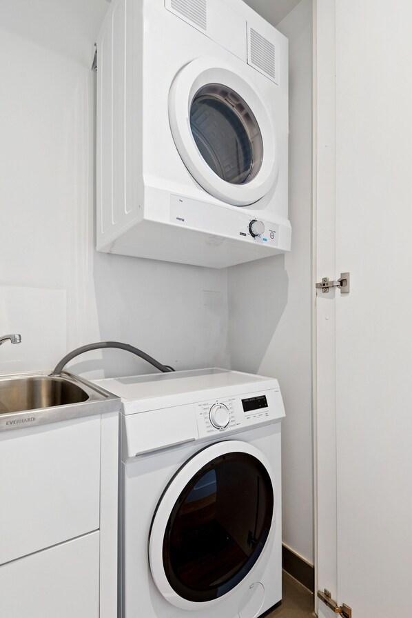 A compact laundry area with a front-loading washer and dryer, a stainless steel sink, and a door to the right. - Bayside Apartment Living Close to the Beach (Melbourne)