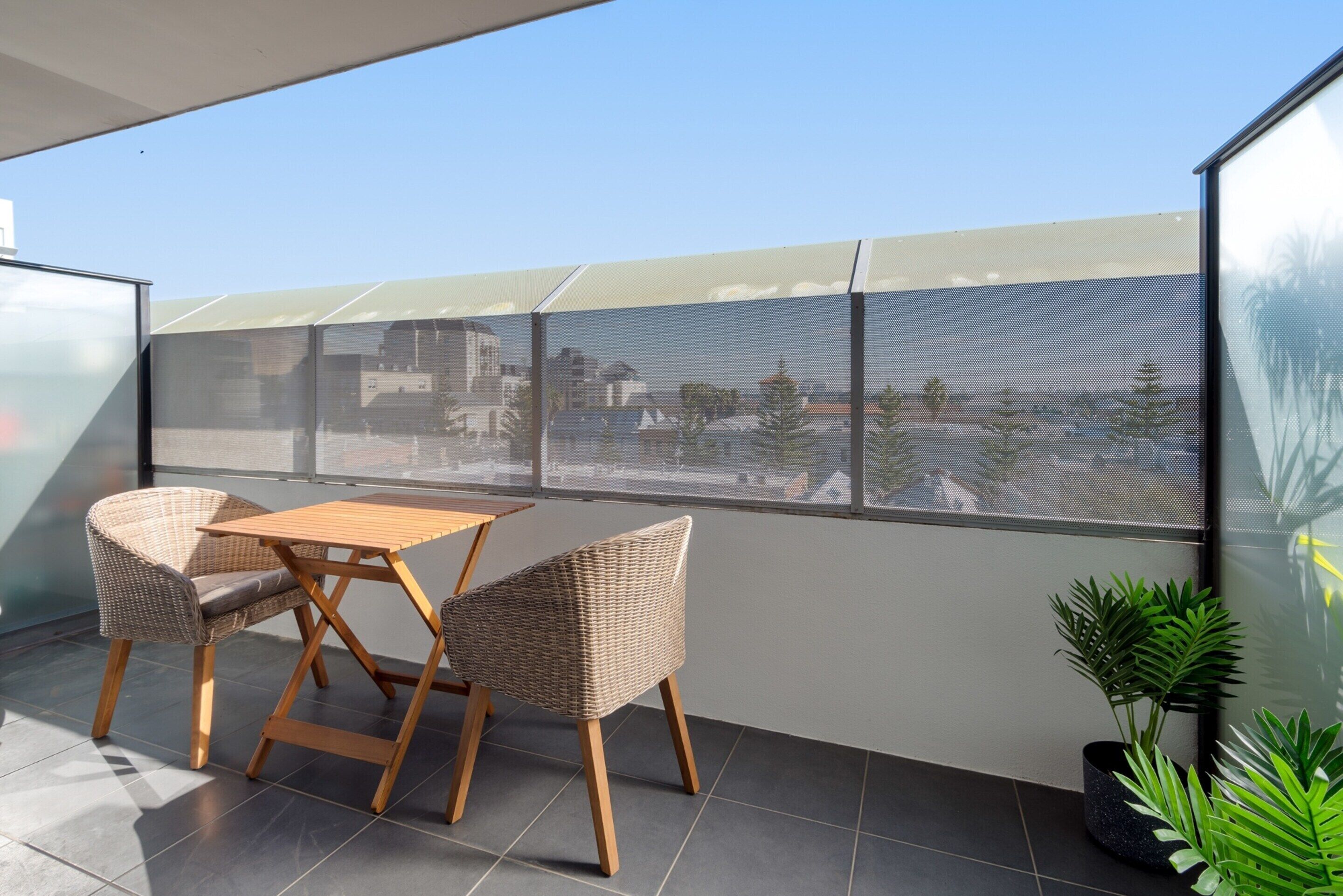 Un balcón con una mesa de madera y dos sillas de mimbre, una planta en maceta y vistas de edificios a través de una malla.