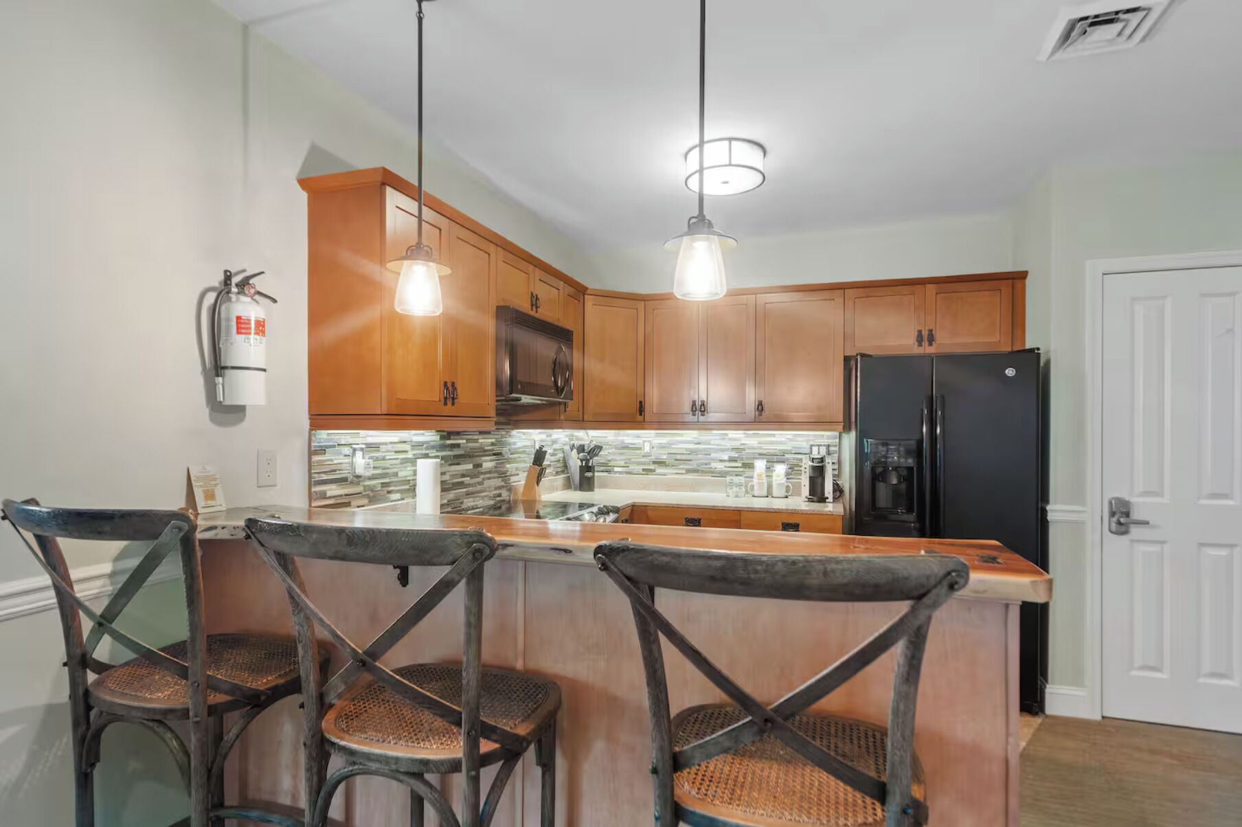 A kitchen with wooden cabinets, a black refrigerator, a coffee maker, and a fire extinguisher. There are three wicker chairs at the island.