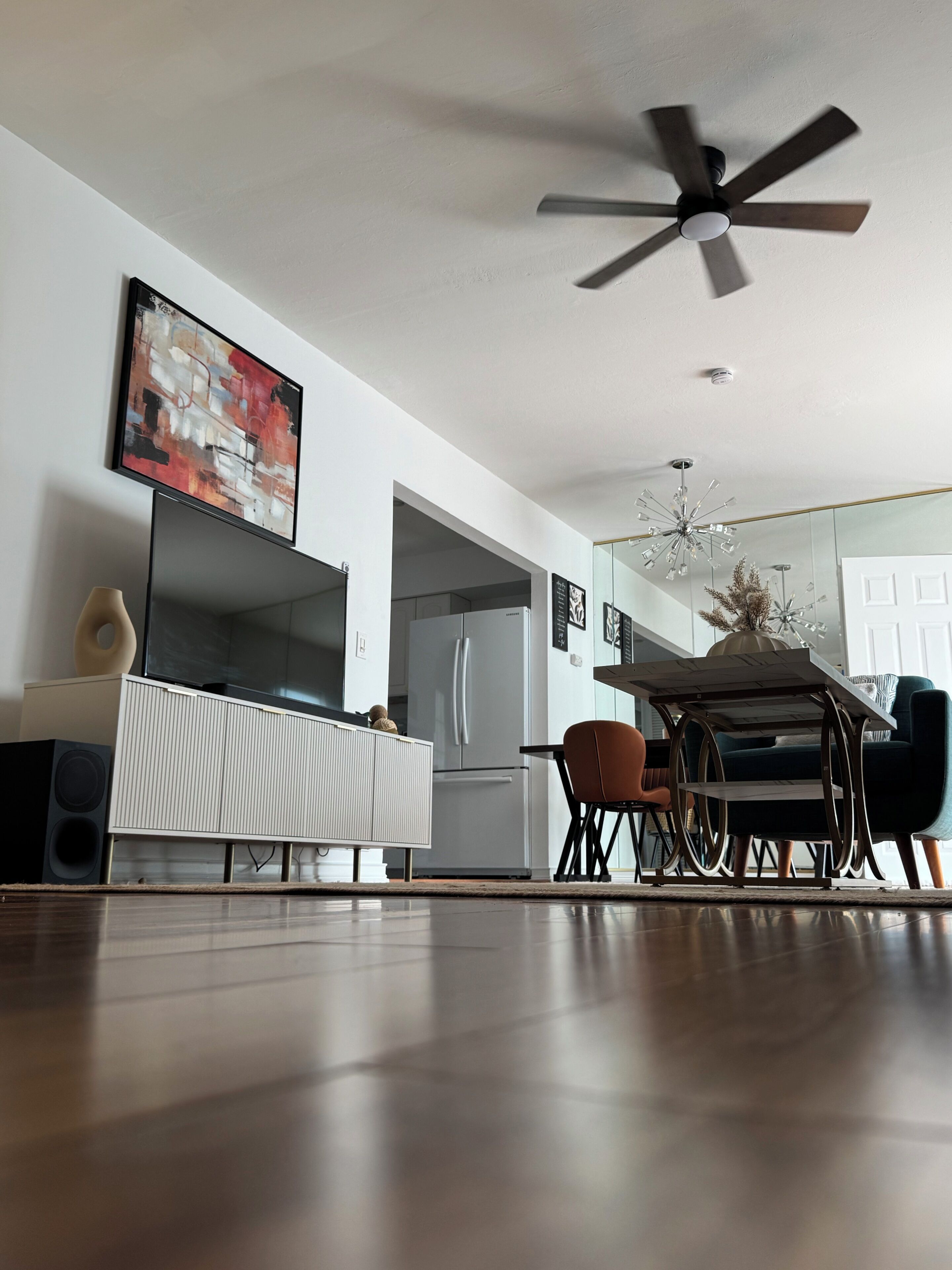 A modern living room with a flat-screen TV, a ceiling fan, a dining area with a table and chairs, and a kitchen with a refrigerator.