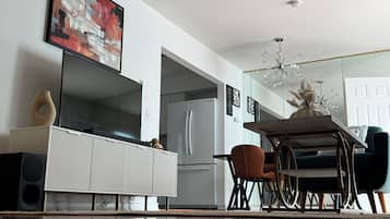 A modern living room with a flat-screen TV, a ceiling fan, a dining area with a table and chairs, and a kitchen with a refrigerator.