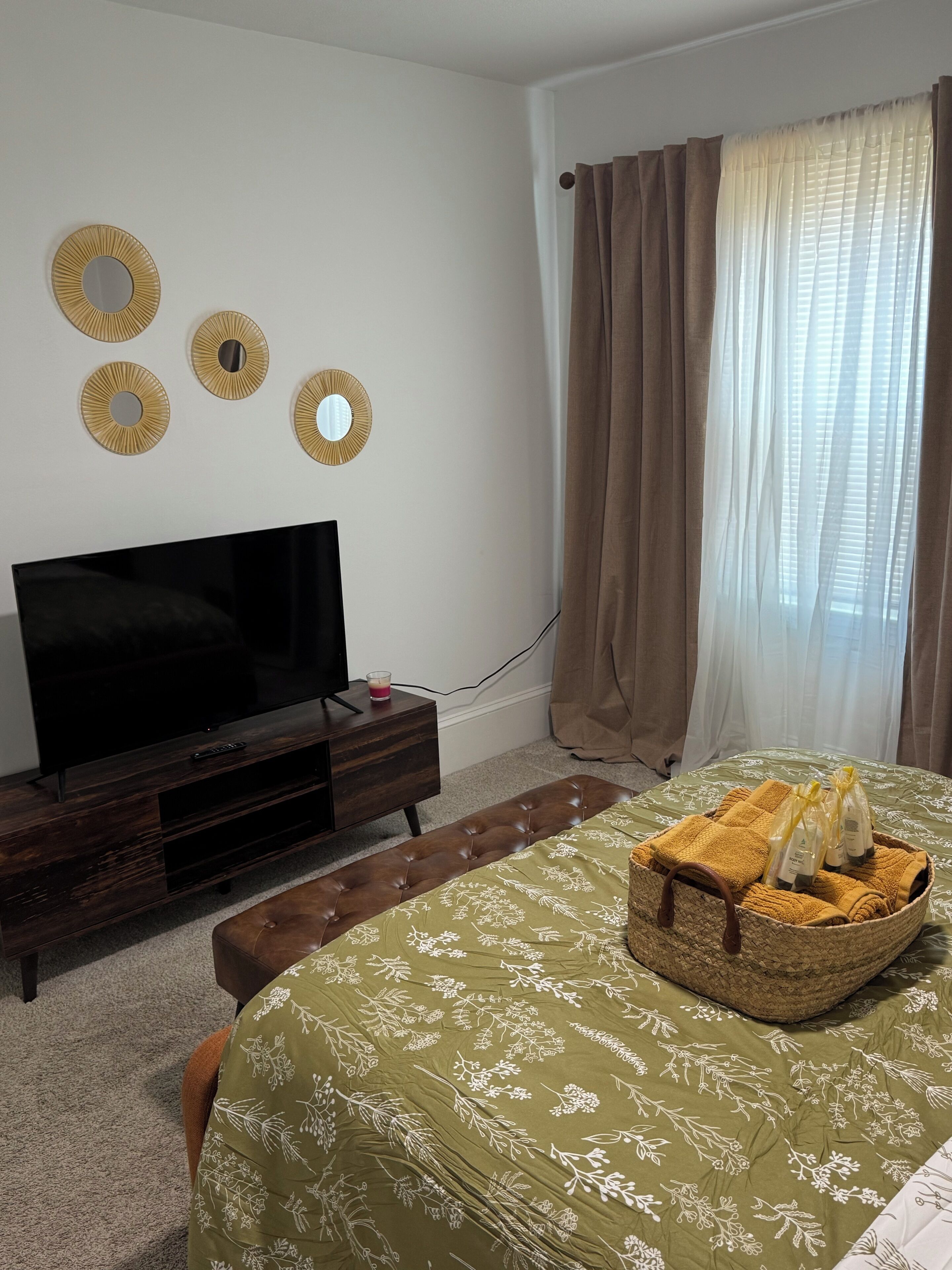 A bedroom with a TV, a bedside table, a bed with a green floral bedspread, a woven basket with toiletries, and a window with curtains.