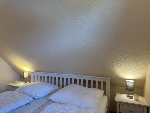 A bedroom with a white bed, two pillows, a nightstand with a lamp, and a small drawer. - Ferienhaus in Burhave (Butjadingen)