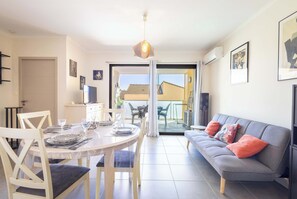 A modern living room with a dining table set for four, a grey sofa with colorful cushions, and a large window offering a view of the outdoors. - Appartement lumineux avec balcon a Valras Plage (Valras-Plage)