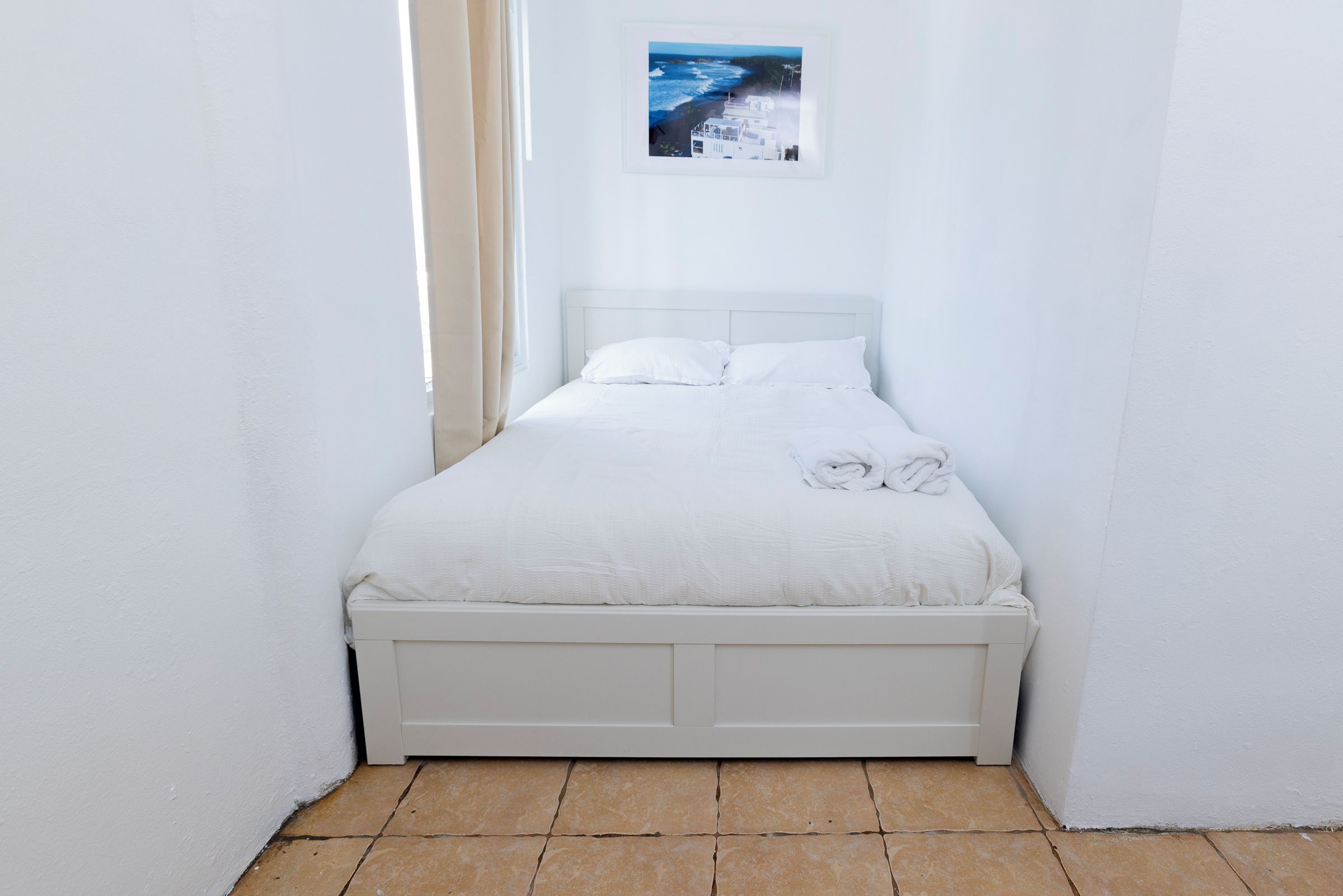 Una cama individual con cabecera y piecera blancos, ropa de cama blanca impecable y una toalla enrollada al lado.