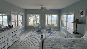A bedroom with a bed, two chairs, a dresser, and a nightstand. There are large windows with a view of the water. - Bayfront home with private pier & dock- near Fairhope & Magnolia Springs (Foley)