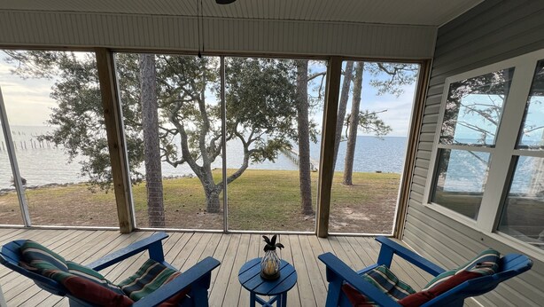 A covered porch with two blue chairs and a small table, overlooking a grassy area with trees and a body of water. - Bayfront home with private pier & dock- near Fairhope & Magnolia Springs (Foley)