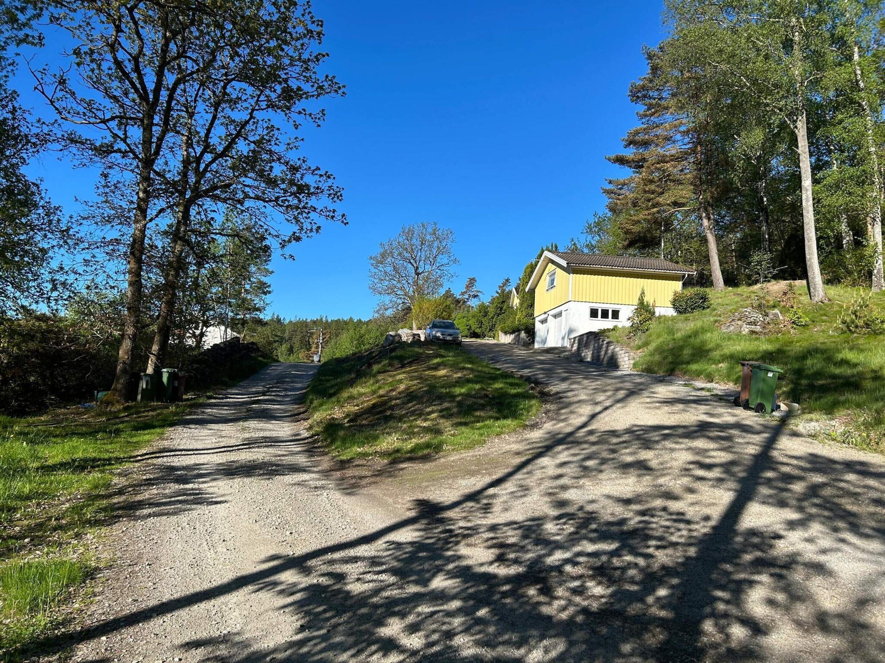 A gravel road leading to a yellow house with a garage, surrounded by trees and greenery.