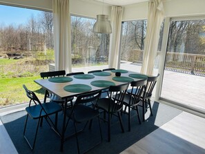 Un espace repas comprenant une table et des chaises, de grandes fenêtres et une vue sur une terrasse et des arbres.