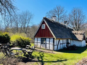 Une maison traditionnelle aux couleurs rouge et blanche, avec un toit de chaume, entourée d’une cour verdoyante et d’arbres.