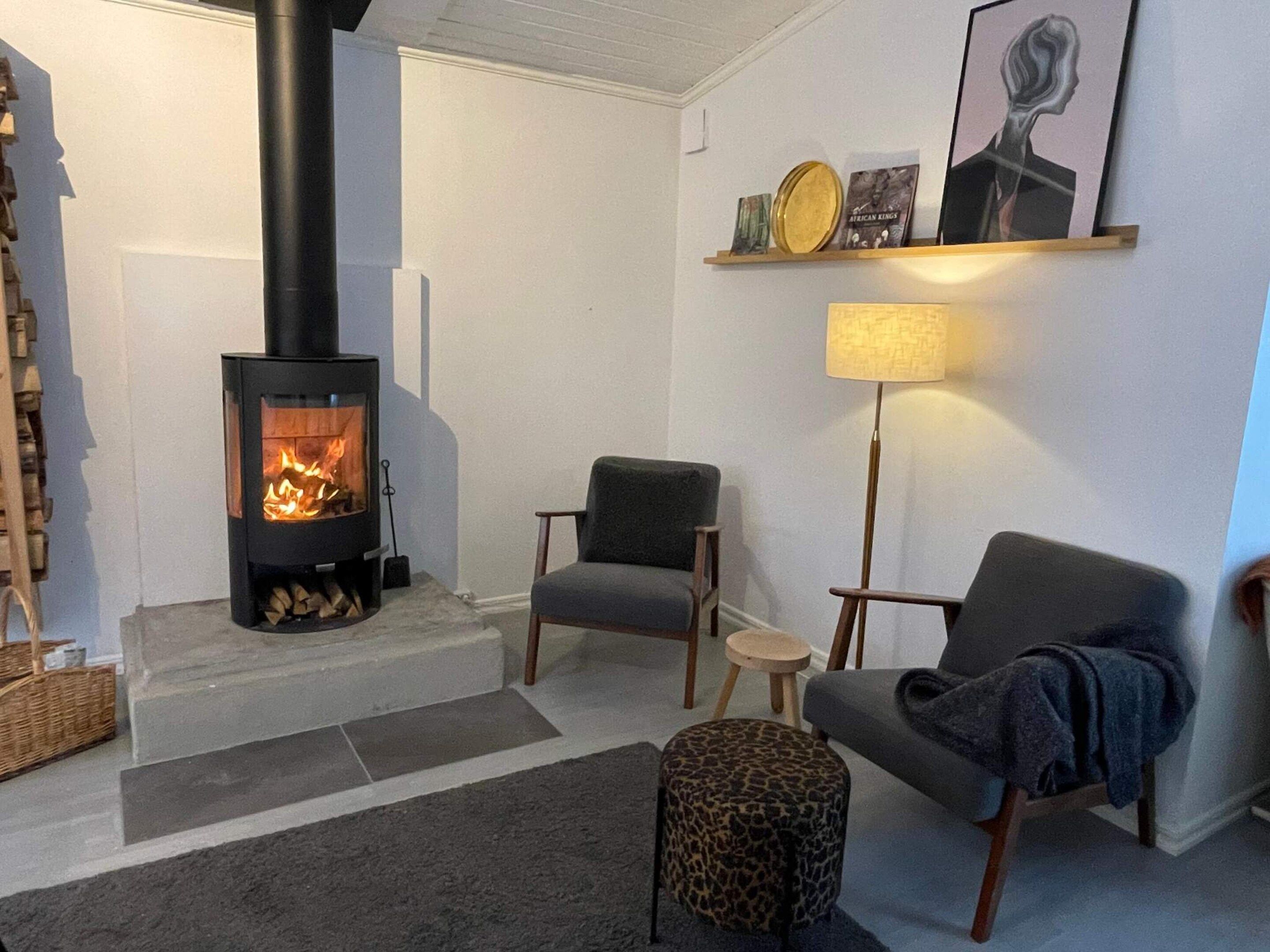 Una sala de estar con un estufa de leña negra, dos sillones grises, un otoman con estampado de leopardo y una lámpara.