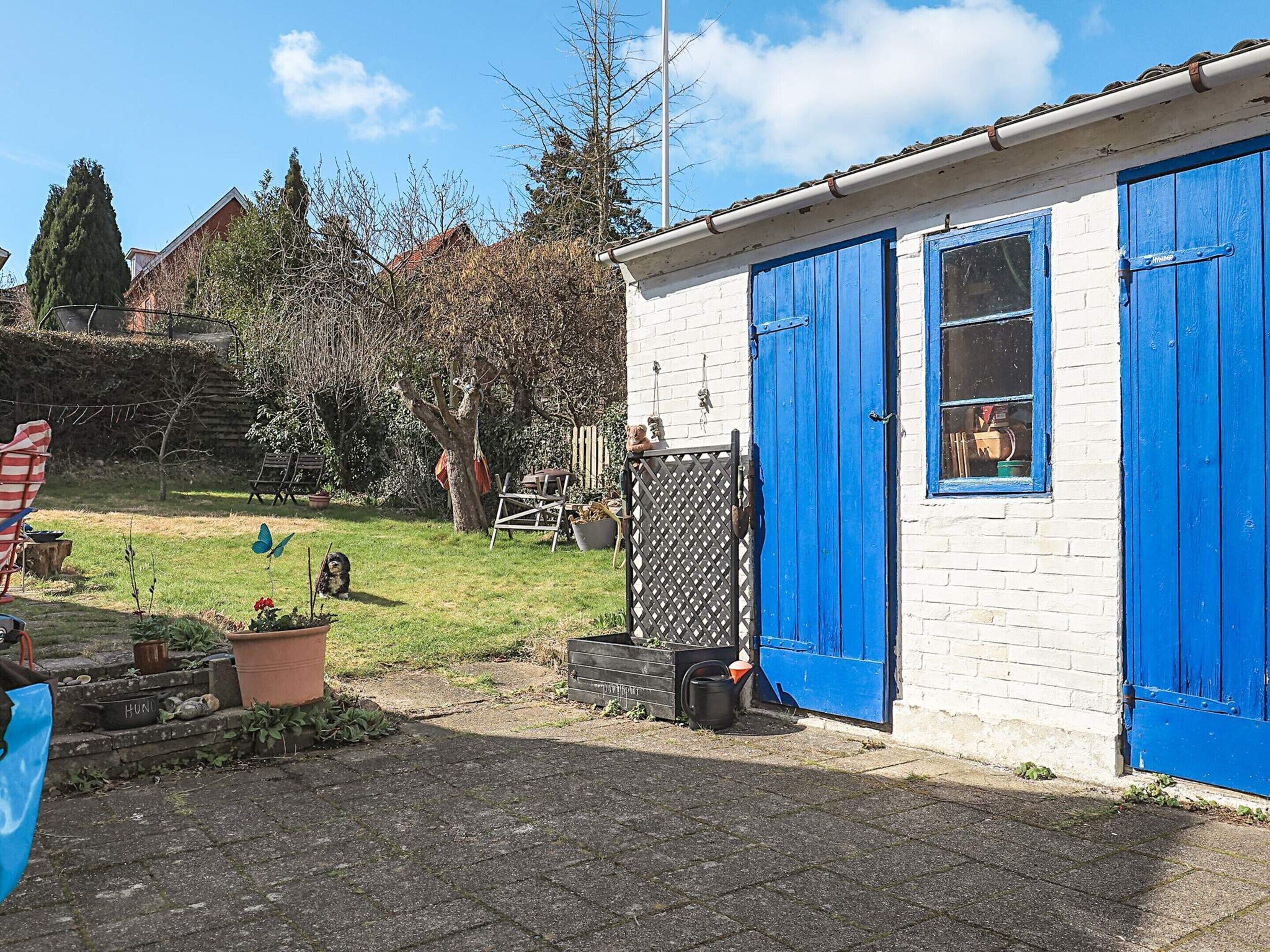 A small white building with blue doors and windows, surrounded by a garden with potted plants and a lawn.