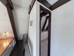 Um corredor estreito com chão de madeira, uma cadeira preta e uma escrivaninha com abajur.