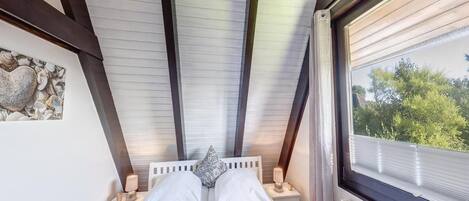 Quarto com teto inclinado, cama com roupa branca, mesa de cabeceira com luminária e janela com vista para o verde.