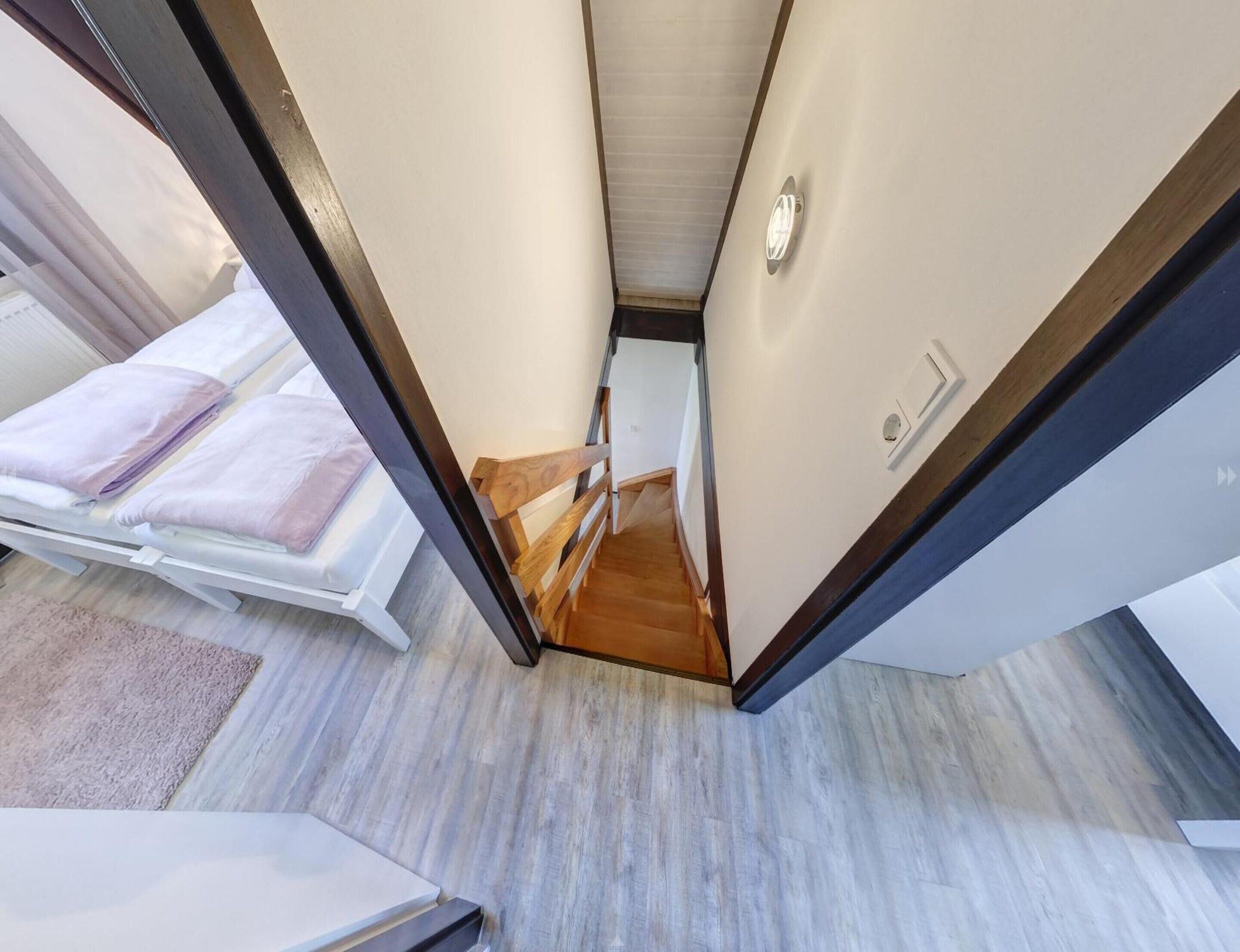 Un couloir étroit avec un escalier en bois menant à une chambre à coucher équipée d’un lit et d’une tête de lit blanche.