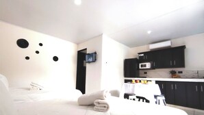 A modern hotel room with a neatly made bed, a small kitchen area with dark cabinets and a microwave, and a wall-mounted TV. - Studio/Pool/WIFI-Full kitchen-A/C-Large Beach-Sunset-Surf***. (Playa Grande)