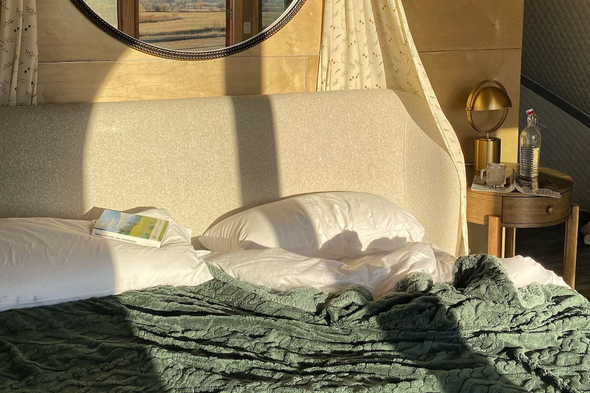 Un dormitorio con cama, una mesita de noche, lámpara, una botella y una ventana con cortinas.