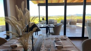 Un coin repas avec une table dressée pour deux personnes, un vase contenant des plantes séchées et une vue sur un vignoble par de grandes fenêtres.