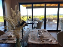 Un comedor con una mesa dispuesta para dos, un florero con plantas secas y vistas a un viñedo a través de amplios ventanales.