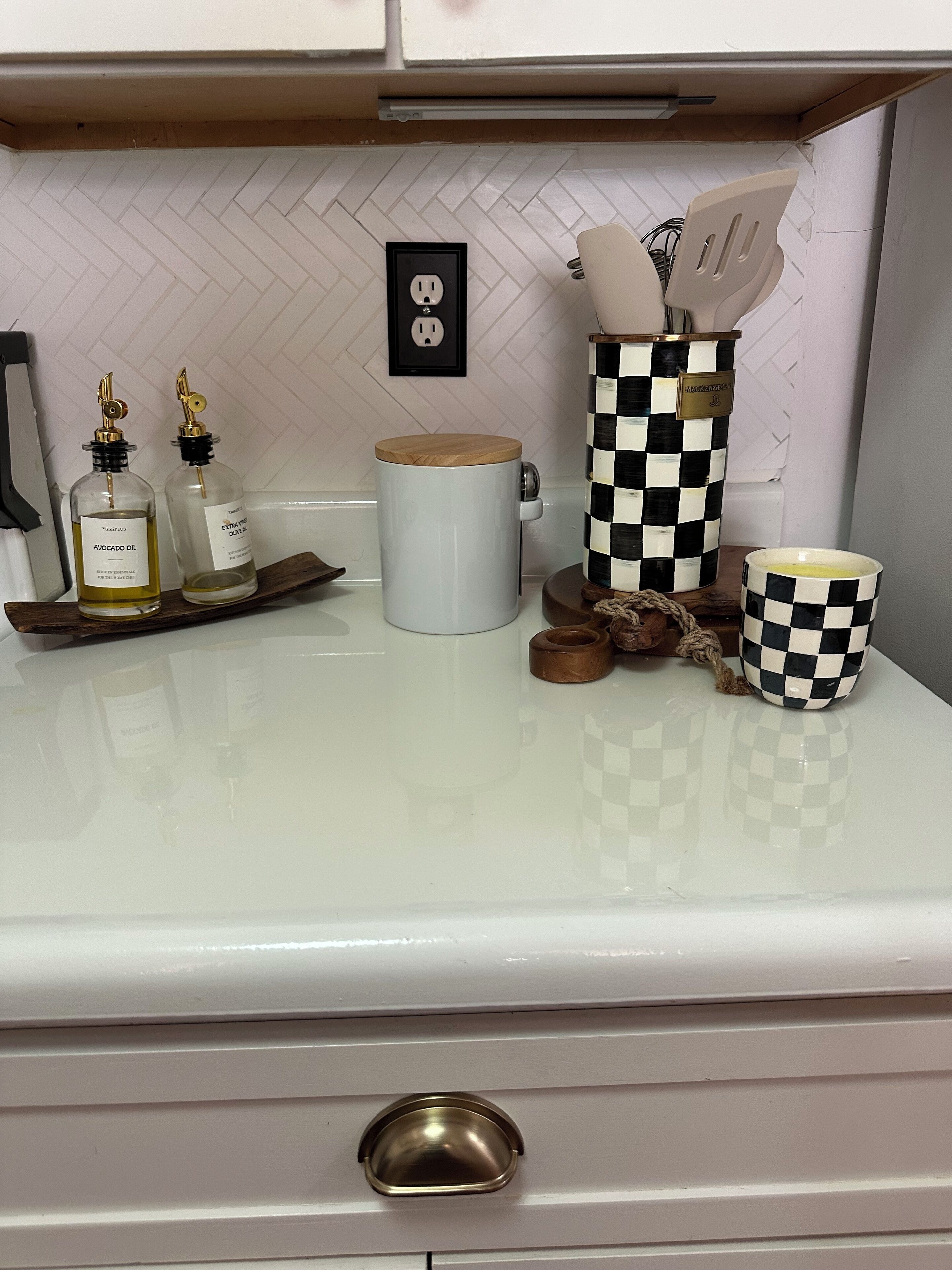 A kitchen counter with two bottles of oil, a white mug, a checkered container, and a checkered mug.