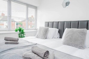 Una cama bien hecha con ropa de cama y almohadas grises, una ventana con persianas y una planta verde en el alféizar.
