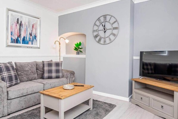 Una sala de estar con un sofá gris, una mesa de centro, un reloj de pared, un televisor y un cuadro en la pared.