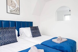 Un dormitorio con una tapicería azul en la cabecera, ropa de cama blanca y almohadas estampadas.