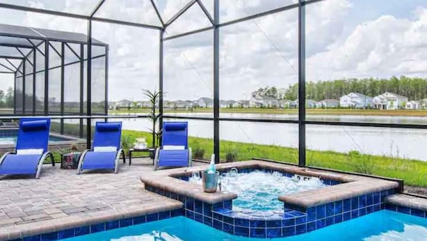 Piscina con hidromasaje y sillas de descanso, rodeada por un recinto de vidrio con vistas a un lago y casas.