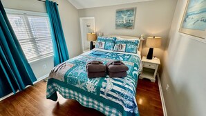 Una cama bien hecha con una colcha azul y blanca con motivos marinos, dos toallas sobre la cama y un cuadro de veleros en la pared.