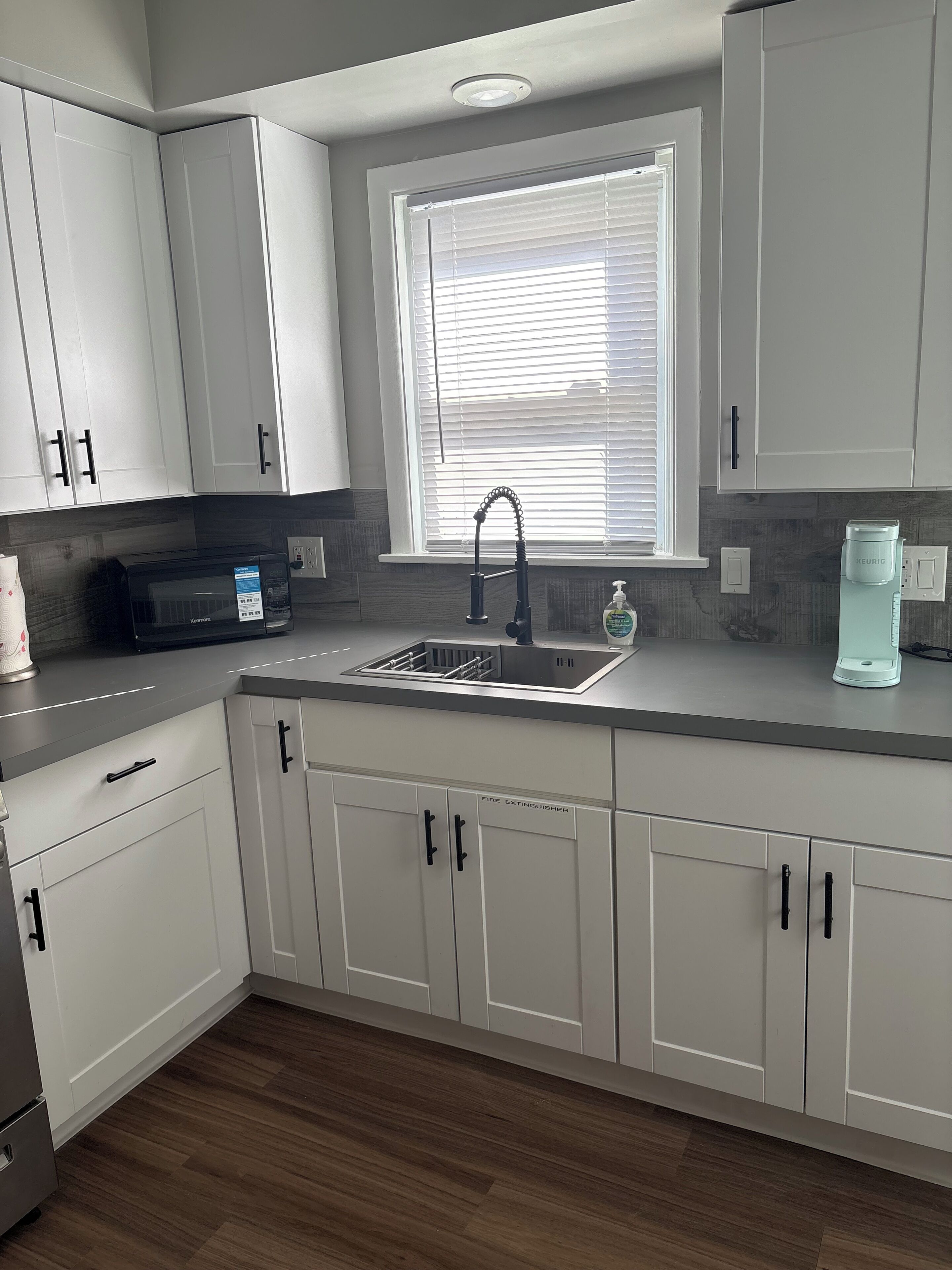 A modern kitchen with white cabinets, a stainless steel sink, a microwave, and a Keurig coffee maker.